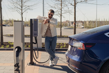 A stylish adult male checks his watch as he waits by his electric car, which is charging at a...