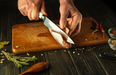 The cook hands use a knife to cut frozen hake fish on a wooden cutting board. National fish dish prepared in a restaurant according to a unique recipe
