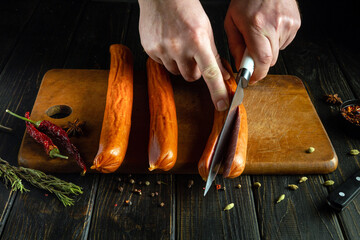 A cook with a knife in his hand cuts sausage on a kitchen board to prepare a snack. Low key concept...