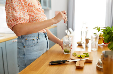 culinary, drinks and people concept - close up of woman with glass and spoon making lime mojito cocktail at home kitchen