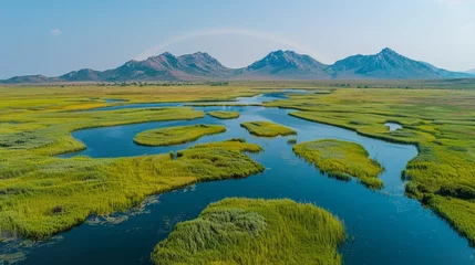 Foto op Canvas   A bird's-eye perspective of a serene river encircled by verdant grasslands, framed by distant mountains, and graced with a rainbow arching across © Alice