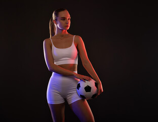 A beautiful athletic blonde girl in white shorts and a top plays football on a black background in neon light.