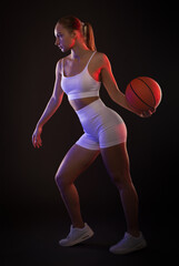 A beautiful athletic blonde girl in white shorts and a top plays basketball on a black background in neon light.