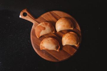 Overhead view of freshly fried empanadas on a round wooden tray on a black table.