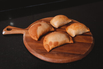 Freshly fried empanadas on top of a round wooden tray, on a black table.