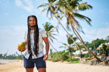 Bipoc man with dreadlocks enjoys fresh coconut water, strolls on sunny tropical beach, relaxed vibe, palm trees backdrop, clear blue sky, embodying travel, leisure and healthy lifestyle for POC.