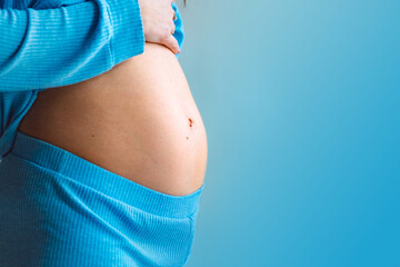 Close-up of young pregnant woman's belly on blue color background. Motherhood and baby expectation concept