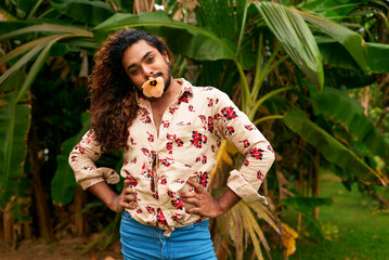 Confident dark-skinned man with floral shirt, blue jeans holds yellow flower in mouth, strikes fashionable pose in garden setting. Cheerful expression, celebrates diversity, LGBTQ pride, summer vibes.