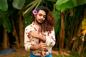 Confident gay man with curly hair, flower in hair stands in tropical garden. BIPOC individual in...