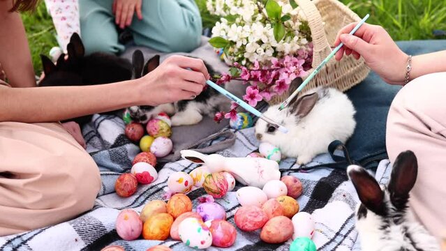 A group of cute little bunnies play in nature near girls painting Easter eggs. Easter bunny and easter eggs concept. Girls touch a rabbit with paint brushes.