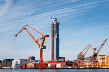 Fototapeta na wymiar Vivid orange cranes stand tall at Gothenburg port against a backdrop of skyscrapers and blue skies, concept of maritime and industry. Gothenburg, Sweden