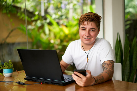 Transgender individual works remotely, multitasking with laptop, smartphone at home office. Smiling, pro, connected, inclusive work environment. Casual biz, wireless tech, body art visible.