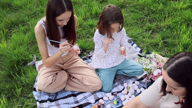 a family of three girls of different ages and European appearance are sitting in the garden on the green grass and are engaged in painting Easter eggs. Easter traditions. Spring holiday.
