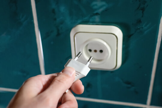A hand holding an electric plug with a socket on the wall in the bathroom