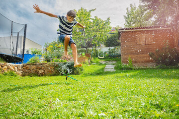 Happy boy illuminated by sunshine hopping over lawn sprinkler - 784033084