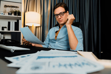 Smart businessman holding paperwork report paragraph dynamic database, looking at pc screen with analyzing global target evaluation value profit company at modern home office at night time. Pecuniary.
