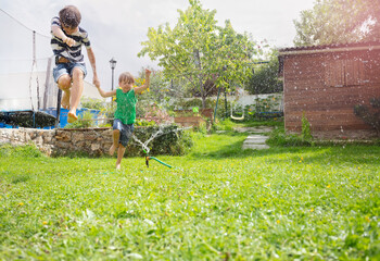 Two boys leap and splash around in sunlit garden with sprinkler - 784034285