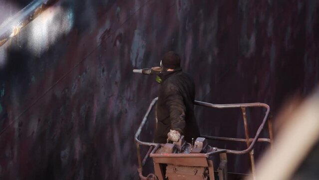 Worker in protective gear uses sandblaster on ship hull in floating dry dock. Industrial paint removal, surface prep, rust elimination, marine maintenance. Shipyard job, vessel refurbishment, repair.