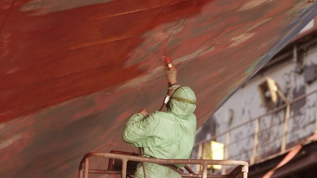 Worker in protective gear applies red paint to ship hull in dry dock. Industrial painting, maintenance against rust, maritime vessel refurbishment process. Shipyard worker, safety equipment in use.