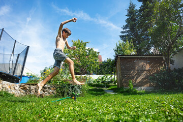 Boy bursts with energy as he vaults a spray of water in backyard - obrazy, fototapety, plakaty