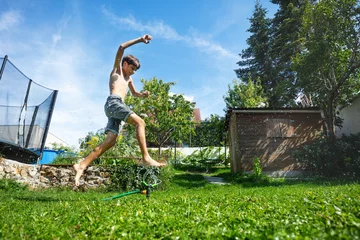 Rucksack Boy bursts with energy as he vaults a spray of water in backyard © Sergey Novikov