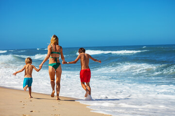 Two boys and a woman in swimwear sprinting joyfully across beach - 784041436