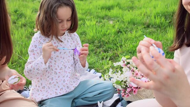 a family of three girls of different ages and European appearance are sitting in the garden on the green grass and are engaged in painting Easter eggs. Easter traditions. Spring holiday.