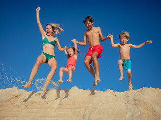 Mother with kids jump into air on sandy beach against blue sky - 784053275