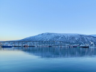 The Tromsdalstinden mountain is reflected in the particularly calm waters of the Tromso harbor on an early January morning, with boats still docked.