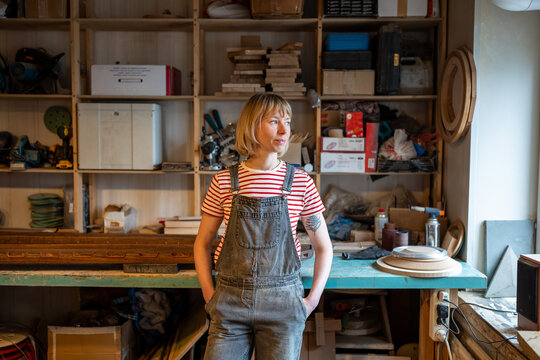 Thoughtful woodworker woman take break in carpentry workshop smiling looking to window pondering creative project. Calm joiner female relaxed standing in artisan room daydreaming, hands in pockets.