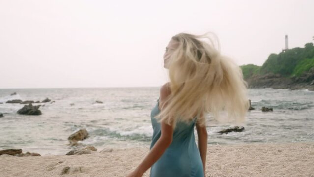 Blonde woman in blue dress walks along tropical beach. POV follows model exploring scenic coast with rocks, lighthouse. Female enjoys summer sea breeze, waves. Casual coastal lifestyle, wanderlust.