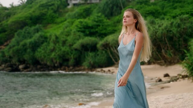Elegant woman strolls along beach, blue dress flowing. Blonde hair catches light, serene coastal backdrop. Leisurely walk by sea, luxury escape. Sunset, solitude, tropical escape reflected Slowmo