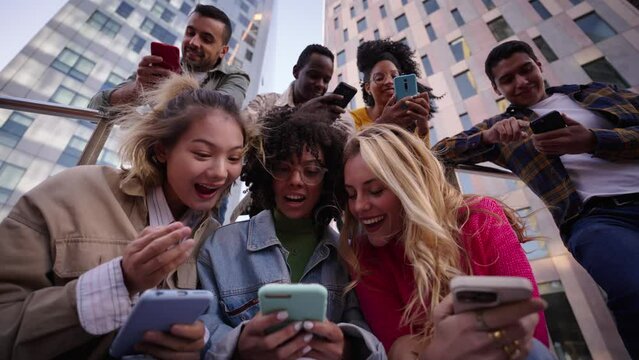 Group of young people watching something at phone sitting on stairs. Smiling multicultural friends looking social media outside the university campus. Students in their free time.