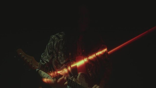 Young long-haired guitarist playing rock music in dark room with strobe light and beam shining from above. Shaky cam shot