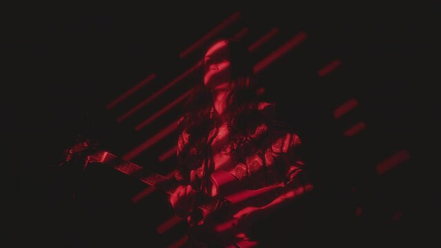 Young groovy indie musician playing the guitar and enjoying the rhythm in dark studio with red light projection