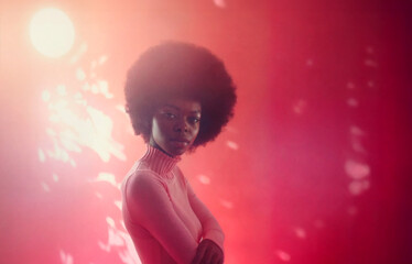 A young African American woman poses confidently with a serious expression against a disco-style pink and red background with a spotlight. She is wearing a high-neck pink sweater and an afro hairstyle