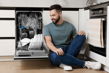 Smiling man sitting near open dishwasher in kitchen