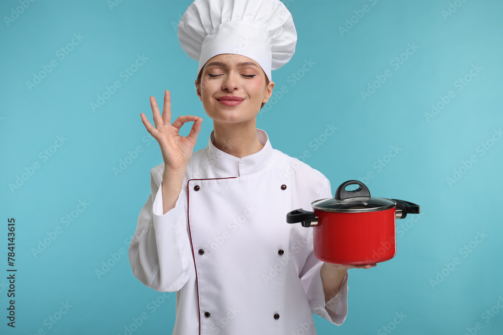 Canvas Prints Happy woman chef in uniform holding cooking pot and showing perfect sign on light blue background