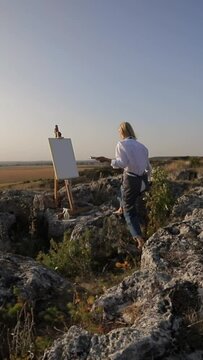 Painting a landscape with sky, water, and buildings on canvas atop a rocky hill