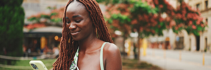 Smiling gorgeous woman with African braids wearing top stands outside on the street and uses mobile...