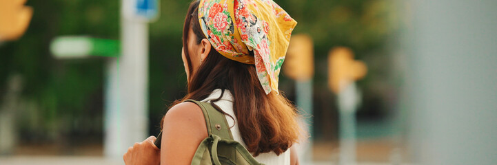 Back view of cute tanned woman with long brown hair in white top and yellow bandana with backpack on her shoulders is walking along pedestrian crossing, Panorama - obrazy, fototapety, plakaty
