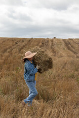 Working woman transporting a hay bale. She is walking sideways in a field of cut wheat. The sky is cloudy. She is dressed in work clothes: denim, gloves, and a sun hat