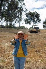 A woman on her cultivated plot lifting both thumbs up as a sign of approval, with hay bales and a tractor in the background