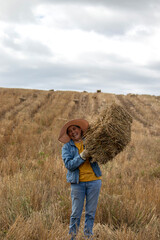 A smiling mature Latina woman dressed as a farmer in jeans lifting a bale of hay on a sunny afternoon with clouds