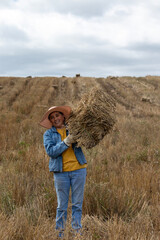 Senior woman carrying a hay bale on her shoulder, dressed in agricultural attire and wearing protective gloves