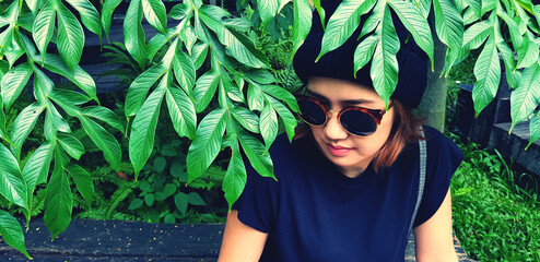 Asian woman wearing sunglass and wool hat sitting on wooden bench under tree with green plant...