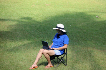 Asian woman in straw beach hat doing online work with computer on folding chair with green grass...