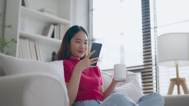 Attractive smiling young Asian woman using mobile phone while sitting on the sofa at home