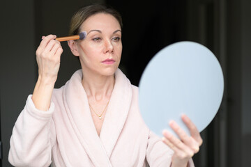 Blonde woman applying makeup with a brush and looking into a mirror