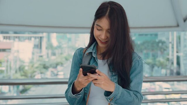 Young asian woman smiling, using mobile smart phone outdoor. Happy beautiful female wearing jeans jacket and holding smartphone at public city. Online technology and communication concept